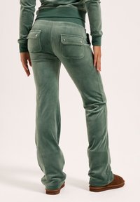 Samtgrüne, ausgestellte Hosen mit einer glatten Textur, versehen mit zwei Gesäßtaschen mit Metallakzenten. Kombiniert mit braunen Schuhen.