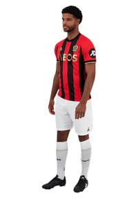 Maillot de football rayé rouge et noir avec le logo "INEOS", shorts blancs, chaussures noires et chaussettes blanches montantes à accents dorés.