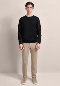 Ein junger Mann steht da, trägt einen schwarzen langärmligen Pullover, beigefarbene Hosen, die am Knöchel umgeschlagen sind, und beigefarbene Sneakers vor einem schlichten Hintergrund.