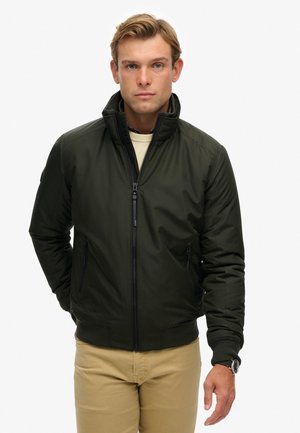 Dark green bomber jacket with a zip front, two side pockets, ribbed cuffs, and collar design. Worn over a light sweater, neutral background.