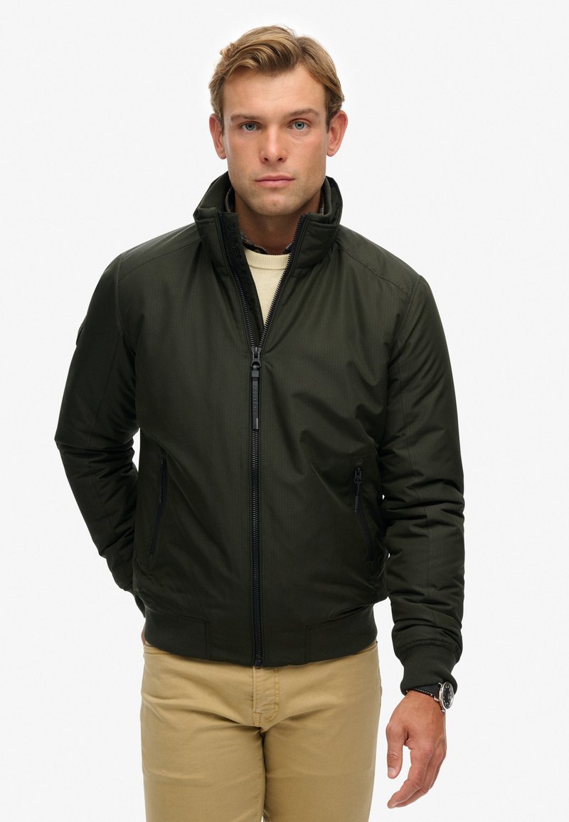 Dunkelgrüne Bomberjacke mit Reißverschluss, zwei seitlichen Taschen, gerippten Bündchen und Kragen. Getragen über einem hellen Pullover, neutraler Hintergrund.