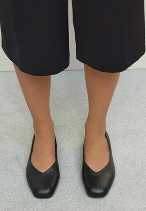 Schwarze Lederflats mit einem spitzen V-förmigen Ausschnitt an der Vorderseite, glatter Textur, leichtem Glanz und einem niedrigen, quadratischen Absatz.