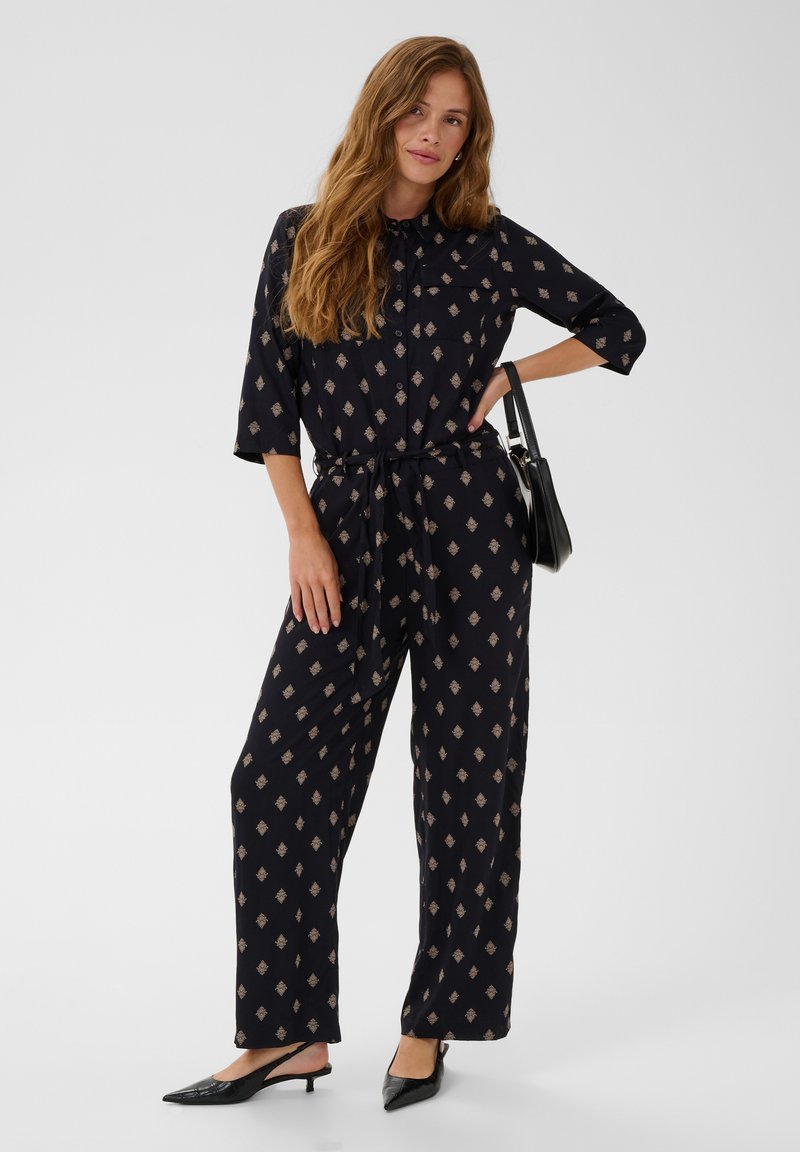 Femme aux longs cheveux portant une combinaison noire à motifs, des chaussures pointues noires et un sac à épaule noir sur un fond uni.