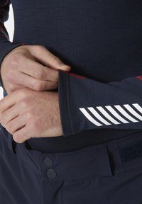 Marineblauwe langemouwen shirt van gestructureerde stof, met rode stiksel en witte gestreepte accenten op de manchetten, gecombineerd met donkere broek en een klittenbanddetail.