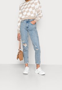 Suéter recortado en argyle en color beige claro y blanco, jeans azul claro desgastados, zapatillas blancas y un pequeño bolso marrón claro con correa para el hombro.