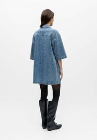 Femme aux cheveux bruns portant une robe-chemise ample en denim bleu, des collants noirs et des bottes noires montantes, de dos sur fond blanc.