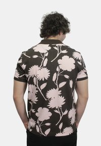 Polo shirt marrone con grandi motivi floreali rosa, maniche corte e colletto. Il tessuto appare liscio con una leggera lucentezza.