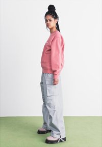 Bomulds sweatshirt i dæmpet pink, parret med grå cargo bukser med lommer. Båret med kraftige, lyserøde klip klip. Neutral baggrund.