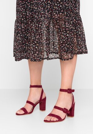 Beine in burgunderfarbenen Wildleder-Sandaletten mit hohen Absätzen und Knöchelriemen, unter einem schwarzen, transparenten Midirock mit Blumenmuster.