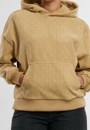 Beige textured hooded sweatshirt with a front pocket worn by a person with hands tucked inside the pocket.