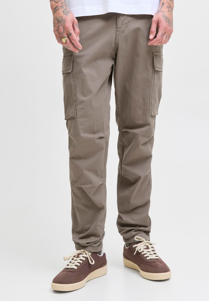 Hombre que lleva pantalones cargo marrones y zapatillas marrones con cordones blancos, de pie frente a un fondo liso.