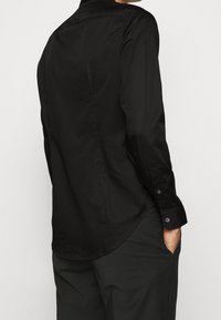 Camisetta nera a maniche lunghe realizzata in tessuto morbido, con orlo curvo, polsini con bottoni e colletto classico, con una vista posteriore mostrata.