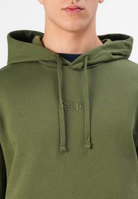 Olive green hoodie with drawstrings, embroidered GAP logo on chest, soft fabric, and a relaxed fit. Textured surface with a smooth finish.