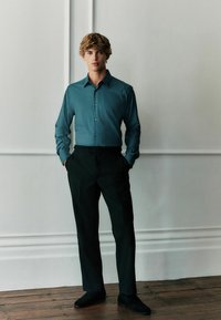 Tyrkysová košela s button-up dizajnom, skombinovaná s čiernymi strihanými nohavicami. Model stojí pred jednoduchou stenou na drevenej podlahe.