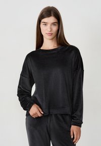 Giovane donna con capelli castani lisci che indossa un top lungo a maniche larghe in velluto nero e pantaloni abbinati, in piedi su uno sfondo bianco uniforme.