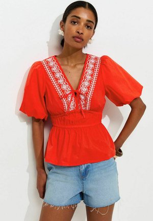 Mujer vistiendo una blusa roja brillante con bordado floral blanco y shorts vaqueros azules, posando con la mano en la cadera contra un fondo liso.