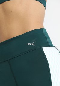 Leggings sportivi di colore verde scuro con un logo argento, caratterizzati da un motivo texturizzato di colore blu chiaro lungo il lato e un design liscio e aderente.