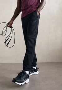 Moški v črnih hlačah EA7, s črnimi in belimi supergami Nike, drži črno vrvico za poskoke in stoji na gladki površini ob bež steni.