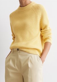 Persona che indossa un maglione di lana giallo con le maniche arrotolate, infilato in pantaloni beige a vita alta, con le mani in tasca, su uno sfondo bianco.