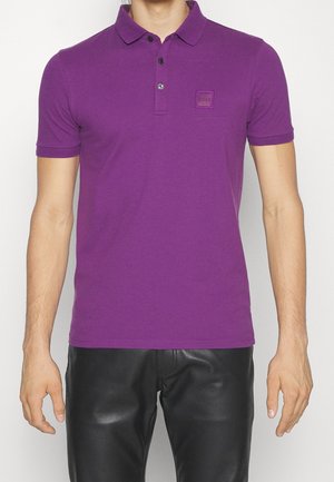 Mann trägt ein eng anliegendes lila Poloshirt mit kurzen Ärmeln und einem kleinen quadratischen Logoaufnäher auf der linken Brust, kombiniert mit schwarzen Lederhosen.