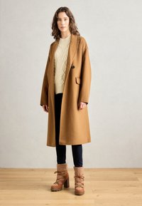 Mujer con un abrigo largo color camel, suéter de punto color crema, pantalones oscuros y botas de invierno con tacón y cordones color beige sobre un suelo de madera.