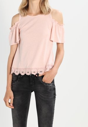 Vrouw die een lichtroze cold-shoulder top met kanten zoom draagt en zwarte gescheurde spijkerbroek, staand met één hand in haar zak.