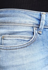 Jeans de denim azul claro con un diseño simple, costuras visibles y un bolsillo frontal. La tela tiene una textura suave con áreas desgastadas.