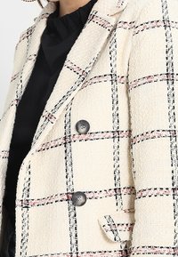 Femme portant un blazer texturé à carreaux crème avec des lignes noires et rouges, sur un haut noir à col montant.