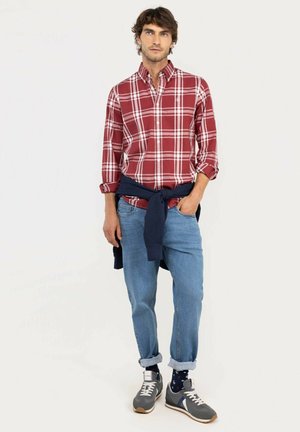 Hombre con camisa de cuadros rojos, vaqueros azules con puños, zapatillas grises y jersey azul marino atado alrededor de la cintura, de pie contra un fondo blanco.