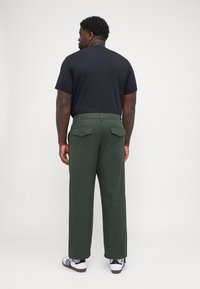 Chemise noire à manches courtes associée à un pantalon large vert avec des poches arrière, portée avec des baskets blanches et noires.