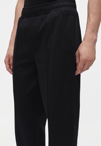 Gros plan d’une personne portant un pantalon noir à taille élastique avec poches latérales, debout, mains détendues le long du corps, sur fond blanc.