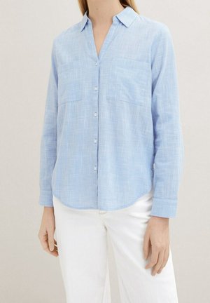 Vrouw draagt een lichtblauw geruit overhemd met knopen en twee borstzakken, en een witte broek, staand tegen een effen lichte achtergrond.