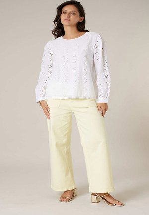 Femme portant un chemisier blanc à œillets à manches longues avec épaules bouffantes, un pantalon large crème et des sandales à talons blocs dorés, debout contre un fond beige.