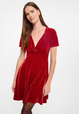 Rotes Samtkleid mit tiefem V-Ausschnitt und kurzen Ärmeln. Ausgestellter Rock mit glatter Textur und sanftem Glanz. Kurz über dem Knie.