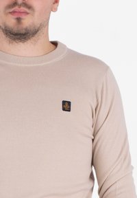 Suéter de punto acanalado beige con cuello redondo y una pequeña etiqueta negra con detalles en dorado. Diseño simple, textura suave y forma ajustada.