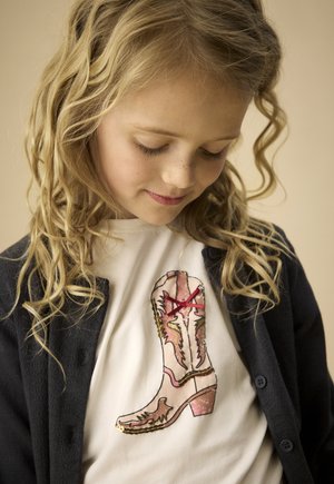 Maglietta bianca con grafica di stivale da cowboy rosa e oro con lacci rossi. Il bambino indossa un cardigan blu navy sopra la maglietta.
