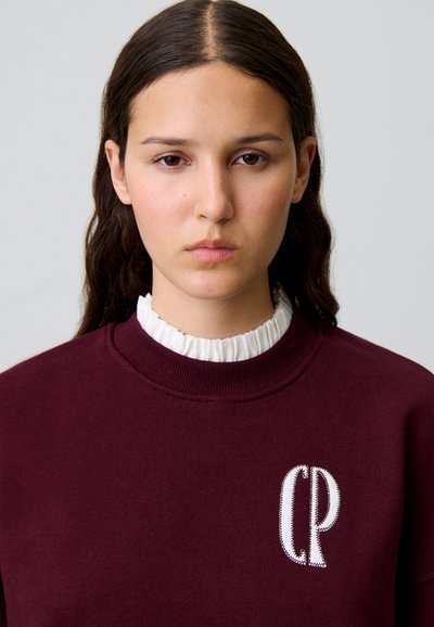 Sudadera burdeos con cuello redondo y logo "CP" bordado en blanco, superpuesta sobre un cuello rizado blanco. Textura suave.