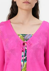 Cardigan rosa a coste con scollatura a V e chiusura con nodo frontale, indossato sopra un top a fantasia floreale con tonalità vivaci di verde e viola.