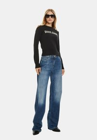 Maglione nero a maglia con logo "GUESS JEANS", abbinato a jeans blu a gamba larga. Modella che indossa occhiali da sole neri e stivaletti con tacco.