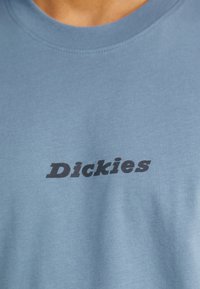 Světle modré bavlněné tričko s černým logem "Dickies" umístěným na hrudi. Tkanina má hladkou texturu s klasickým kulatým výstřihem.