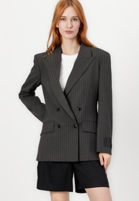 Mørkegrå dobbelspent blazer med smale hvite pinstriper, spiss slag, to frontlommer og kontrasterende mansjettdetalj. Kombinert med svarte shorts.
