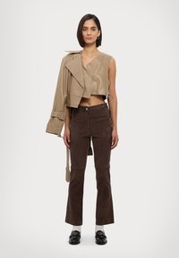 Kürzer beiges Lederjackett über die linke Schulter gelegt; ärmelloses, passendes Oberteil; braune Wildleder-Hose mit Schlag; schwarze Loafer mit goldenen Akzenten.