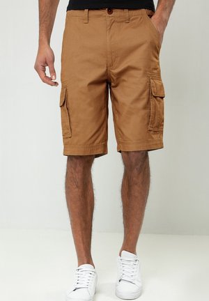 Mand iført brune cargoshorts og hvide sneakers, stående med den ene hånd i lommen mod en ensfarvet baggrund.