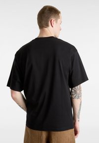 Black cotton t-shirt with short sleeves, featuring a relaxed fit and a plain back design. Visible tattoo on the arm, worn with tan pants.