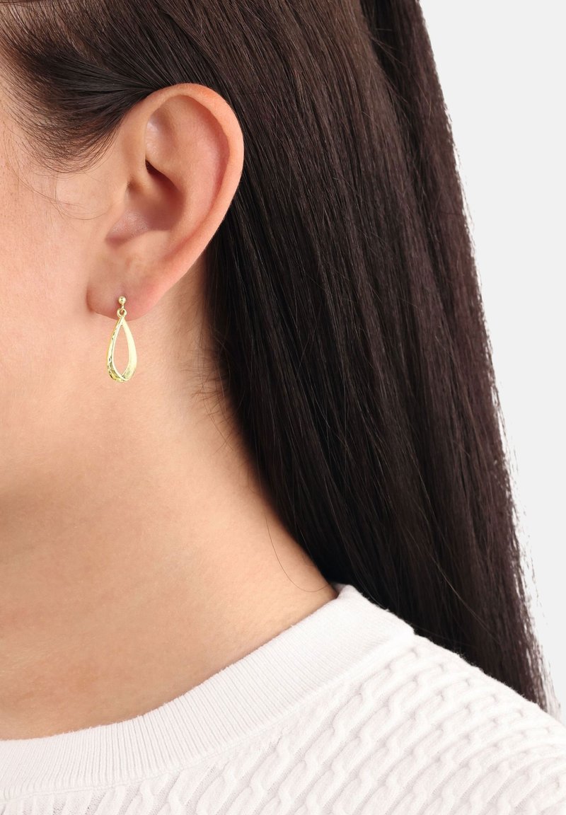 Kvinnas vänstra öra med rakt mörkt hår och en hängande guldöronring i form av en tår, klädd i en vit strukturerad topp.