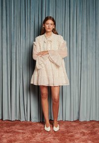 Biele šaty s farebnými bodkami, volánikovými manžetami a kravatovým golierom. Modelka má na sebe biele špicaté topánky. V pozadí sú modré závesy.