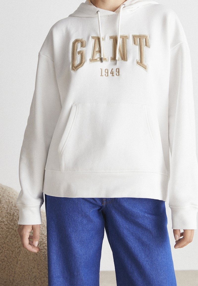 Sudadera blanca con bolsillo frontal, presenta el bordado de "GANT" y "1949" en beige. Incluye cordones ajustables y puños acanalados.