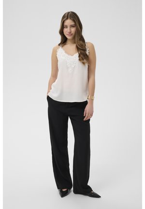 Noor naine seisab taskutes kätega, kandes valget pitsääristusega varrukateta tuba, musti lõdva säärega pükse, musti kingi, kuldset käevõru ja sõrmuseid.