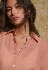 Licht koraalkleurig linnen shirt met een klassieke kraag, voorzien van knoopdetails en een zachte textuur. Draagt oorbellen als accent.