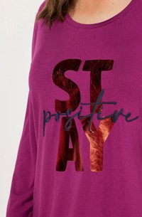 Nahaufnahme eines lila Hemdes mit glänzenden roten Buchstaben "STAY" und schwarzem, geschwungenem Text "positive" über der Brust.